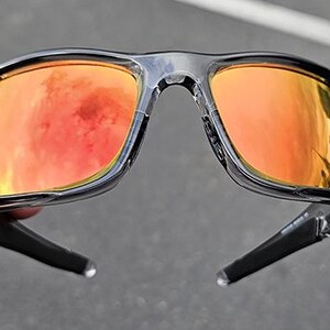 May include: Black sunglasses with orange mirrored lenses. The sunglasses have a glossy black frame and arms. The lenses reflect an orange and red hue, suggesting a bright outdoor setting. The sunglasses are held by a hand.