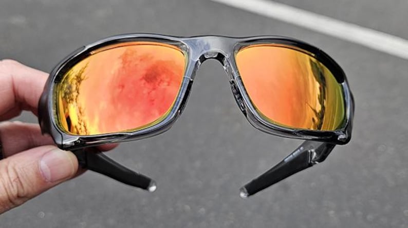 May include: Black sunglasses with orange mirrored lenses. The sunglasses have a glossy black frame and arms. The lenses reflect an orange and red hue, suggesting a bright outdoor setting. The sunglasses are held by a hand.