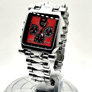May include: A silver-toned Oakley wristwatch with a square red face and black subdials. The watch features a detailed metal band and is displayed on a silver stand. The watch face has the brand name "Oakley" printed on it.