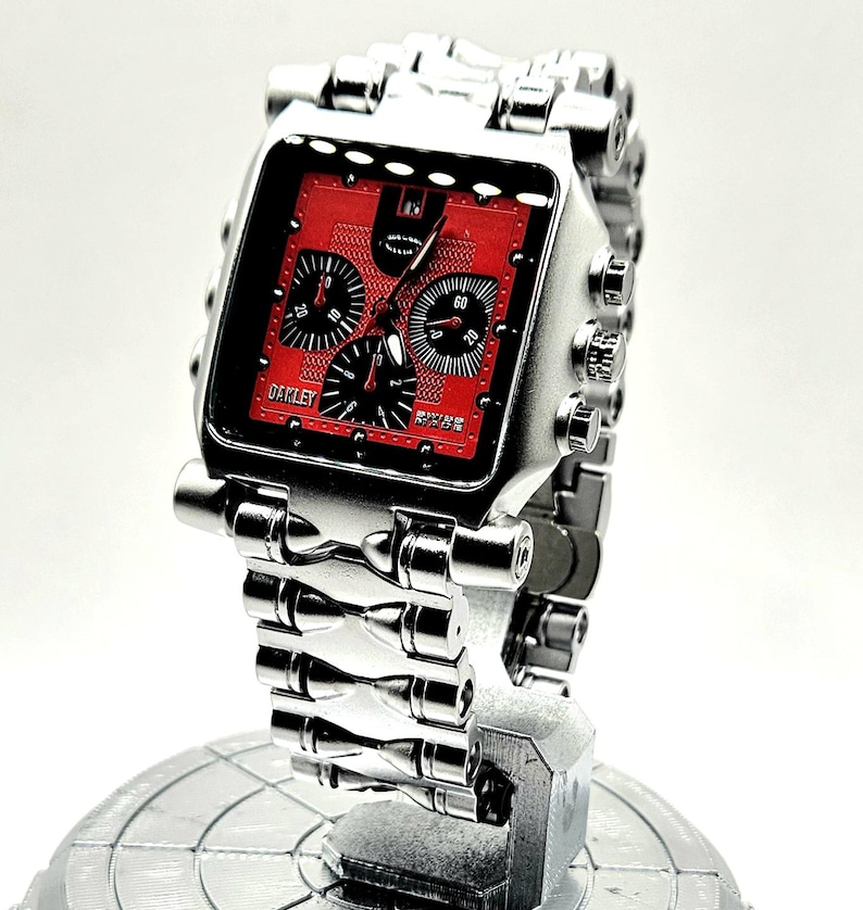 May include: A silver-toned Oakley wristwatch with a square red face and black subdials. The watch features a detailed metal band and is displayed on a silver stand. The watch face has the brand name "Oakley" printed on it.
