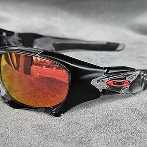 May include: Black sunglasses with orange-tinted lenses. The frames are black with clear accents near the temples. A small red logo is visible on the temple. The lenses reflect the surrounding environment.