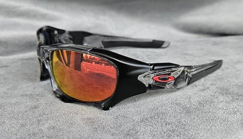 May include: Black sunglasses with orange-tinted lenses. The frames are black with clear accents near the temples. A small red logo is visible on the temple. The lenses reflect the surrounding environment.