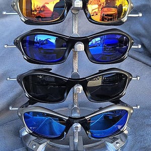 Puede incluir: Exhibici&oacute;n de cuatro pares de gafas de sol con lentes espejadas. El par superior tiene lentes naranjas, el segundo lentes azules, el tercero lentes negros y el inferior lentes azules. Las monturas son negras y plateadas, sobre un soporte met&aacute;lico plateado.