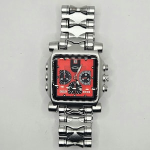 May include: A silver-toned Oakley wristwatch with a square red face. The watch features black subdials and hands, with the Oakley logo visible. The watch band is also silver-toned and has a unique, geometric design.