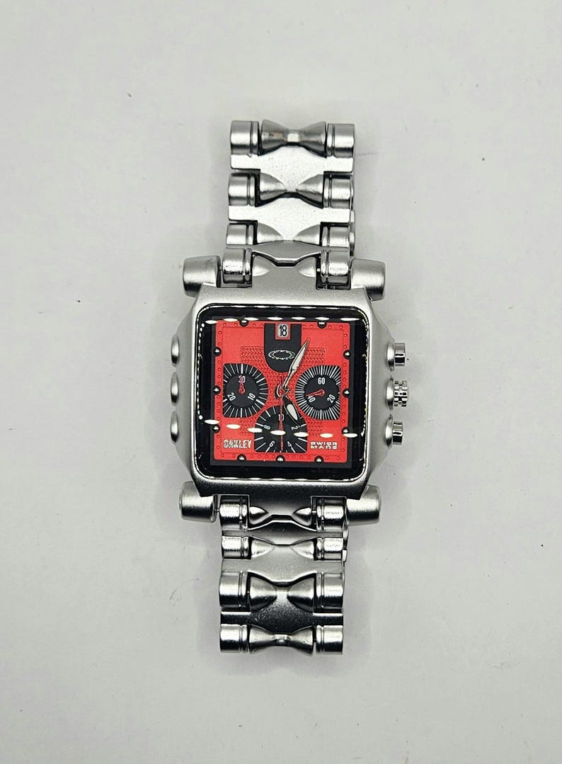 May include: A silver-toned Oakley wristwatch with a square red face. The watch features black subdials and hands, with the Oakley logo visible. The watch band is also silver-toned and has a unique, geometric design.