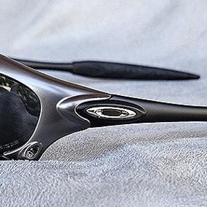 Peut inclure: Une paire de lunettes de soleil gris m&eacute;tallis&eacute; avec des verres fonc&eacute;s. Les lunettes ont un design &eacute;l&eacute;gant et moderne avec des montures incurv&eacute;es et des branches noires. Le logo Oakley est visible sur la monture.