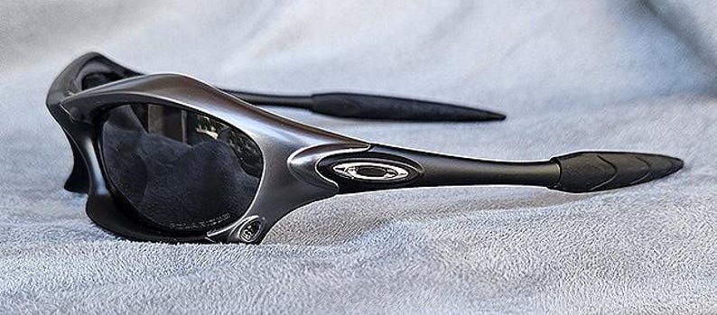 Peut inclure: Une paire de lunettes de soleil gris m&eacute;tallis&eacute; avec des verres fonc&eacute;s. Les lunettes ont un design &eacute;l&eacute;gant et moderne avec des montures incurv&eacute;es et des branches noires. Le logo Oakley est visible sur la monture.