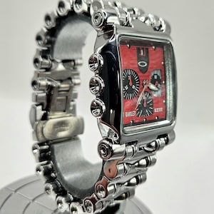 May include: A silver Oakley wristwatch with a rectangular red face and black sub-dials. The watch features a unique, industrial-style band with multiple cylindrical links. The watch is displayed on a silver stand.