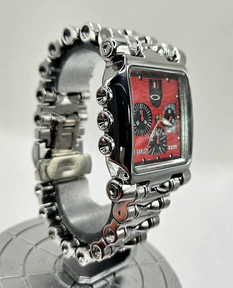 May include: A silver Oakley wristwatch with a rectangular red face and black sub-dials. The watch features a unique, industrial-style band with multiple cylindrical links. The watch is displayed on a silver stand.