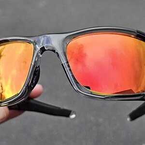 May include: Black and clear-framed sunglasses with orange-red lenses. The sunglasses have a sporty design with a curved frame and temples. The lenses reflect a bright, warm light.