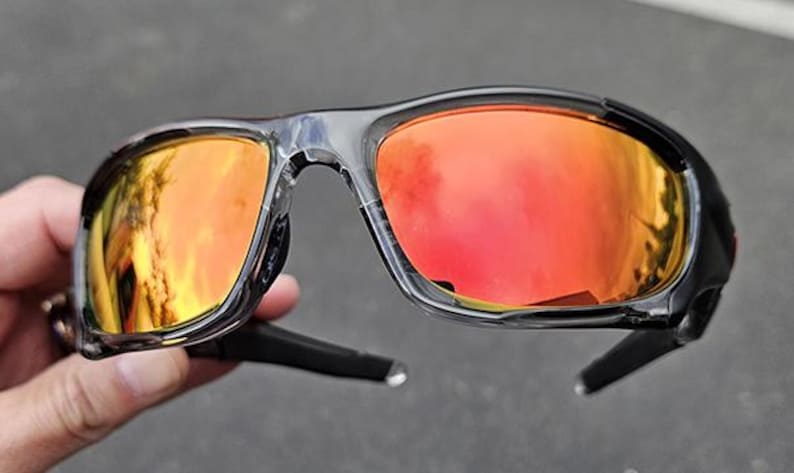 May include: Black and clear-framed sunglasses with orange-red lenses. The sunglasses have a sporty design with a curved frame and temples. The lenses reflect a bright, warm light.