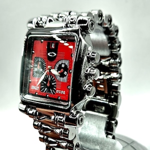 May include: A silver-toned Oakley watch with a rectangular red face. The watch features black subdials and hands, with the Oakley logo visible. The watch band is also silver-toned and has a unique, industrial design.