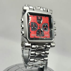 May include: A silver-toned Oakley wristwatch with a square red face. The watch features black subdials and hands, and the brand name is visible on the face. The watchband is also silver-toned.