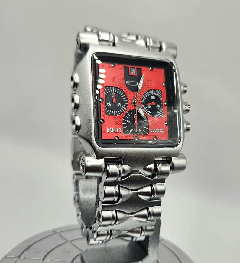 May include: A silver-toned Oakley wristwatch with a square red face. The watch features black subdials and hands, and the brand name is visible on the face. The watchband is also silver-toned.
