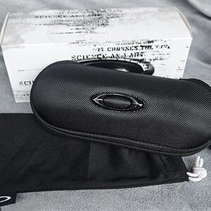 May include: Black zippered sunglasses case with a matching black soft pouch and a white box. The case has a textured surface and a metal emblem. The box has text that reads "SCIENCE AND ART".