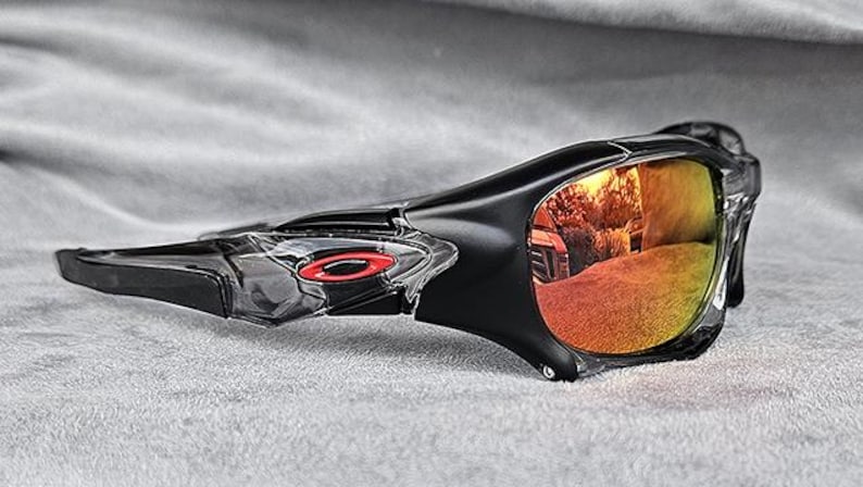 May include: Black sports sunglasses with a wraparound design. The lenses have a gradient of yellow to orange, reflecting the surroundings. A red logo is visible on the frame.