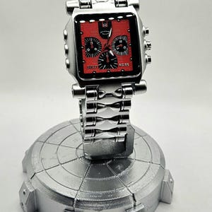 May include: A silver-toned Oakley watch with a square red face and black subdials. The watch is displayed on a silver stand. The watch band and stand have a metallic finish. The watch face has the Oakley logo.