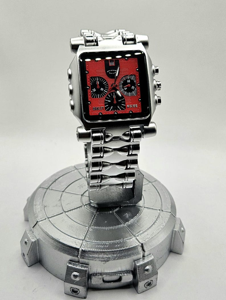 May include: A silver-toned Oakley watch with a square red face and black subdials. The watch is displayed on a silver stand. The watch band and stand have a metallic finish. The watch face has the Oakley logo.