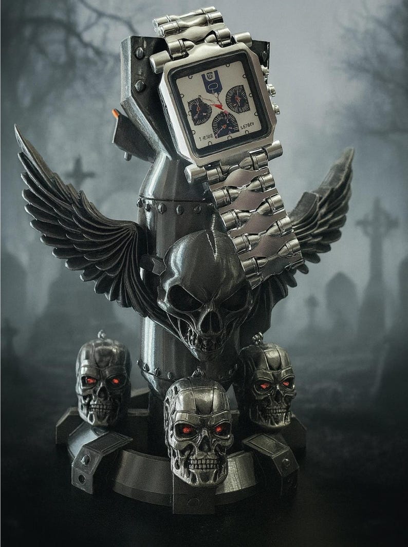 Pode incluir: Um rel&oacute;gio prateado com mostrador quadrado e v&aacute;rios mostradores, repousando sobre um suporte decorativo com uma caveira, asas e caveiras menores com olhos vermelhos. A pulseira do rel&oacute;gio &eacute; de metal.
