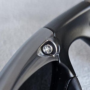 Puede incluir: Primer plano de una secci&oacute;n de gafas de sol gris oscuro. La montura tiene un dise&ntilde;o liso y curvado con un peque&ntilde;o tornillo plateado visible en la bisagra. Las lentes no son visibles.