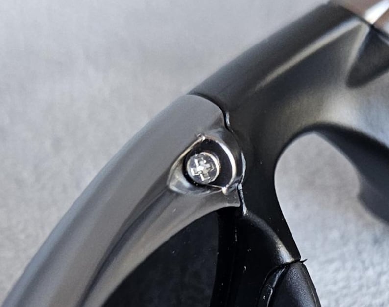 Puede incluir: Primer plano de una secci&oacute;n de gafas de sol gris oscuro. La montura tiene un dise&ntilde;o liso y curvado con un peque&ntilde;o tornillo plateado visible en la bisagra. Las lentes no son visibles.