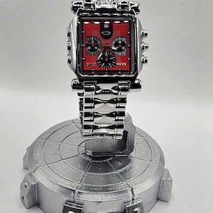 May include: A silver-colored wristwatch with a red face and black subdials is displayed on a silver stand. The watch has a square face with the brand name "Oakley" visible. The watch band is also silver.