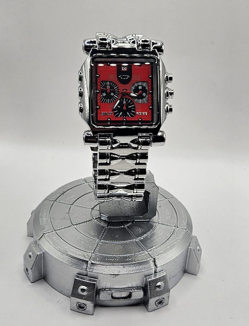 May include: A silver-colored wristwatch with a red face and black subdials is displayed on a silver stand. The watch has a square face with the brand name "Oakley" visible. The watch band is also silver.