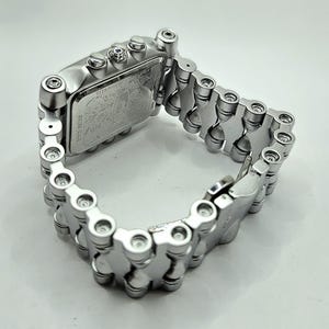 Pode incluir: Um rel&oacute;gio de pulso prateado com mostrador retangular e pulseira em estilo de corrente. O mostrador do rel&oacute;gio apresenta um design detalhado, e a pulseira &eacute; composta por elos arredondados interconectados. O rel&oacute;gio &eacute; mostrado em fundo branco.