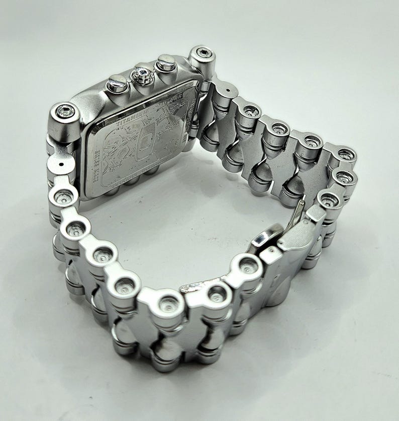 Pode incluir: Um rel&oacute;gio de pulso prateado com mostrador retangular e pulseira em estilo de corrente. O mostrador do rel&oacute;gio apresenta um design detalhado, e a pulseira &eacute; composta por elos arredondados interconectados. O rel&oacute;gio &eacute; mostrado em fundo branco.