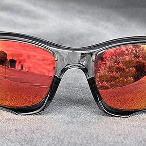 May include: Black-framed sunglasses with mirrored red lenses. The lenses reflect an outdoor scene with a road and trees, creating a vibrant, colorful effect. The sunglasses have a modern, sporty design.