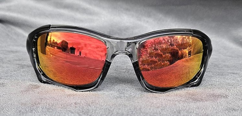 May include: Black-framed sunglasses with mirrored red lenses. The lenses reflect an outdoor scene with a road and trees, creating a vibrant, colorful effect. The sunglasses have a modern, sporty design.