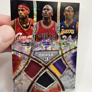 May include: A rectangular sports trading card featuring LeBron James, Michael Jordan, and Kobe Bryant. The card includes three fabric patches, text reading Authentic Patches Triple 3, and NBA SP Game-Used, with a holographic, multi-colored dot pattern.
