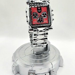May include: A silver-toned wristwatch with a square red face and black subdials. The watch features the brand name "Oakley" and is displayed on a silver stand. The watch band is made of linked metal.
