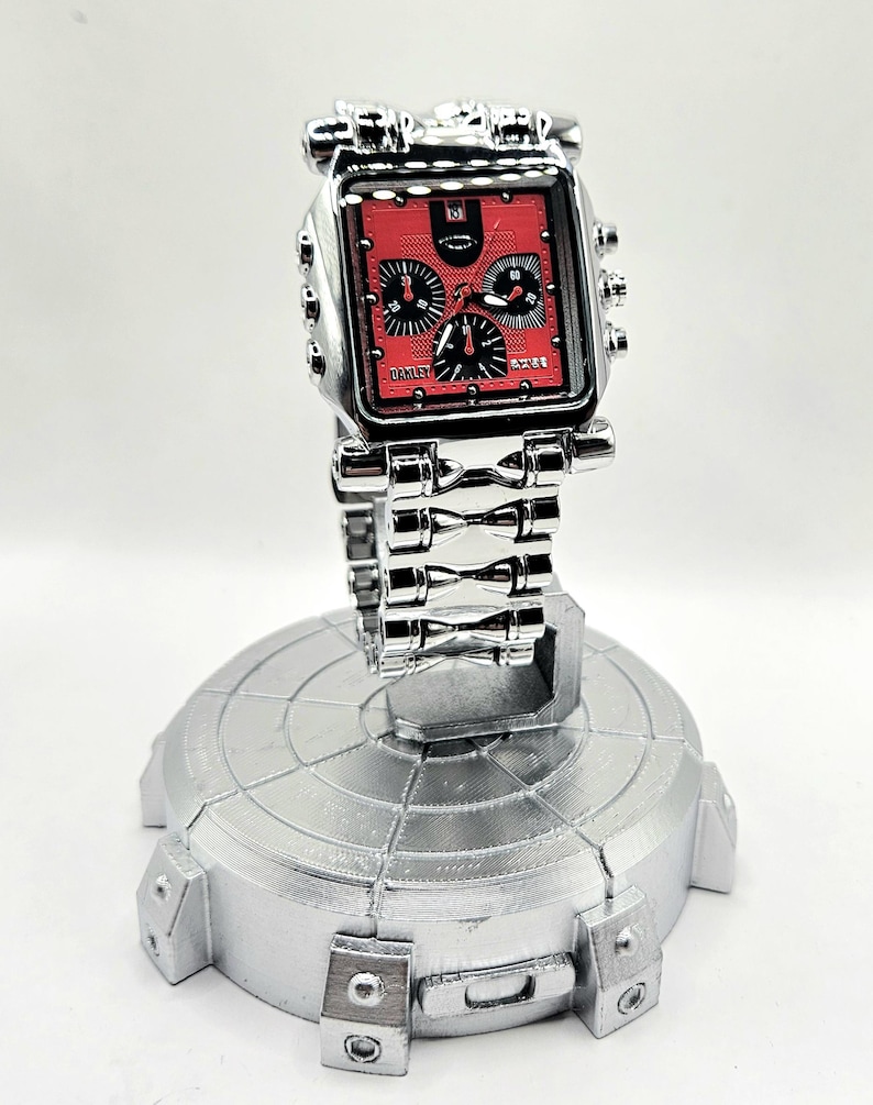 May include: A silver-toned wristwatch with a square red face and black subdials. The watch features the brand name "Oakley" and is displayed on a silver stand. The watch band is made of linked metal.