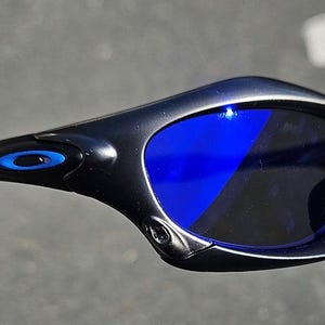 Puede incluir: Gafas de sol negras con lentes azules. Las gafas de sol tienen un dise&ntilde;o elegante y curvado con un logotipo azul en la patilla. Las lentes son de color azul vibrante y las monturas son negras.