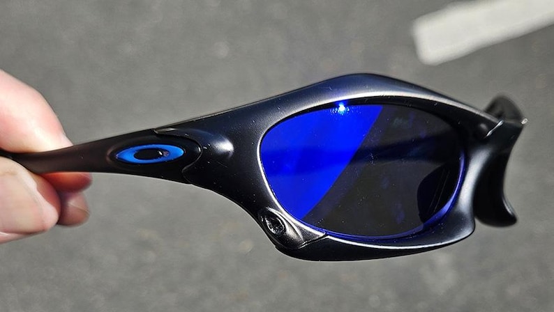 Puede incluir: Gafas de sol negras con lentes azules. Las gafas de sol tienen un dise&ntilde;o elegante y curvado con un logotipo azul en la patilla. Las lentes son de color azul vibrante y las monturas son negras.