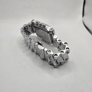 May include: A silver-toned wristwatch with a rectangular face and a chain-link style band. The watch is made of metal and has a clasp closure. The watch is displayed on a white surface.
