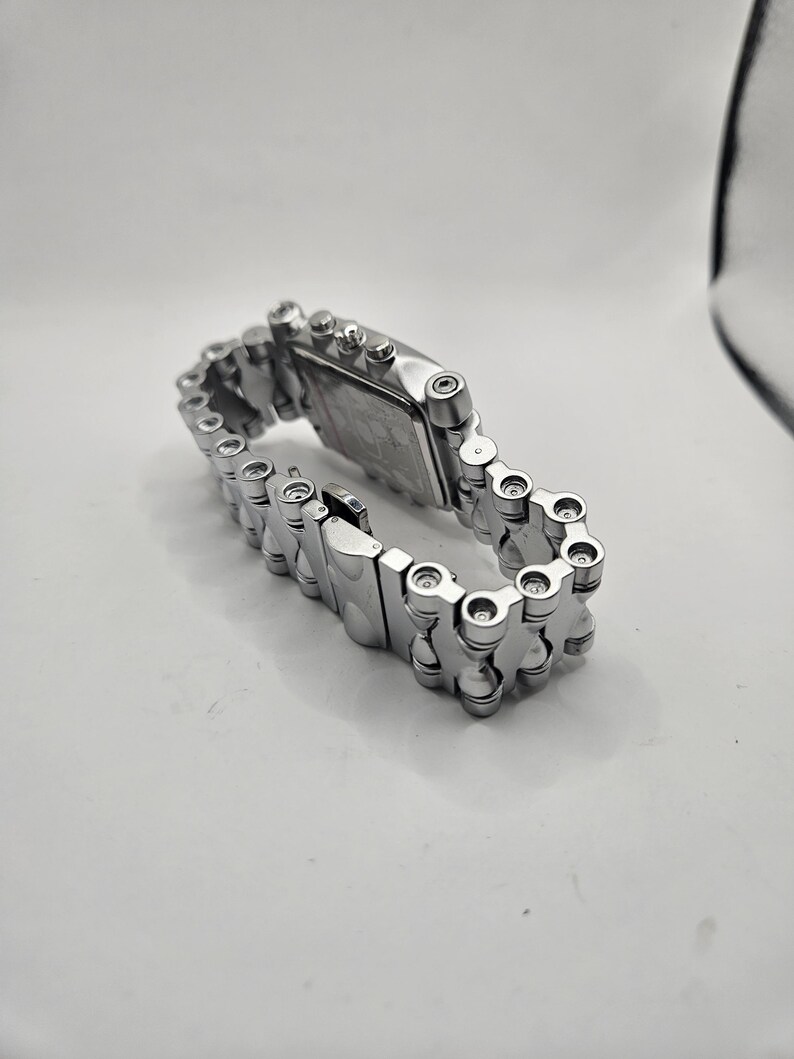 May include: A silver-toned wristwatch with a rectangular face and a chain-link style band. The watch is made of metal and has a clasp closure. The watch is displayed on a white surface.