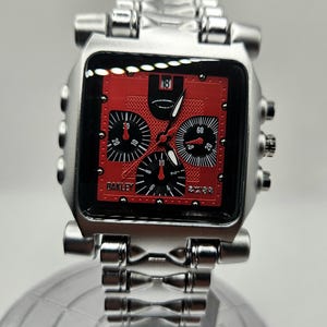 May include: A silver-toned Oakley wristwatch with a square red face. The watch features black subdials and hands, with the brand name "Oakley" visible. The watch band is also silver-toned.