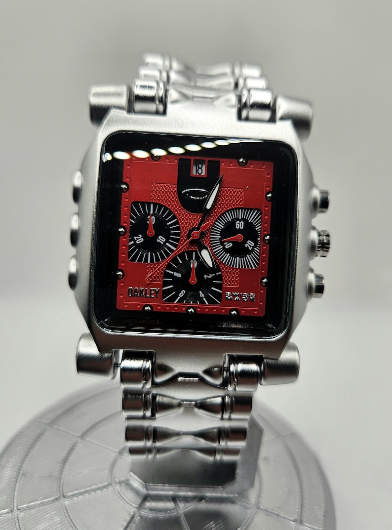 May include: A silver-toned Oakley wristwatch with a square red face. The watch features black subdials and hands, with the brand name "Oakley" visible. The watch band is also silver-toned.
