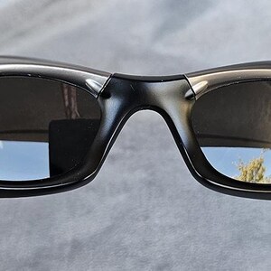 Peut inclure: Lunettes de soleil gris fonc&eacute; avec un design enveloppant incurv&eacute;. Les verres sont sombres, refl&eacute;tant l'environnement. Les montures ont une finition m&eacute;tallique, et le style g&eacute;n&eacute;ral est sportif et moderne. Les lunettes sont tenues par une main.