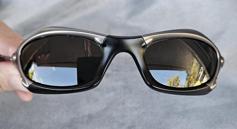 Peut inclure: Lunettes de soleil gris fonc&eacute; avec un design enveloppant incurv&eacute;. Les verres sont sombres, refl&eacute;tant l'environnement. Les montures ont une finition m&eacute;tallique, et le style g&eacute;n&eacute;ral est sportif et moderne. Les lunettes sont tenues par une main.