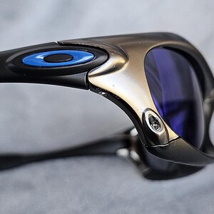 Puede incluir: Primer plano de unas gafas de sol con montura negra y lentes azules. Las gafas de sol tienen un acento met&aacute;lico plateado y negro en la montura con un detalle de c&iacute;rculo azul. Las lentes son de color azul intenso.