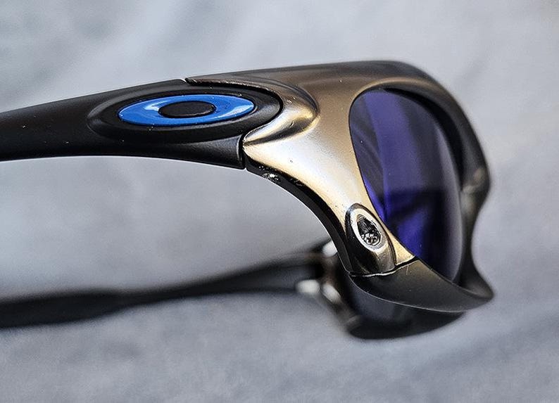 Puede incluir: Primer plano de unas gafas de sol con montura negra y lentes azules. Las gafas de sol tienen un acento met&aacute;lico plateado y negro en la montura con un detalle de c&iacute;rculo azul. Las lentes son de color azul intenso.