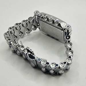 May include: A silver-toned wristwatch with a rectangular face and a chain-link band. The watch face has a detailed design. The band is made of interlocking metal pieces, creating a unique, industrial aesthetic. The watch is shown on a white surface.