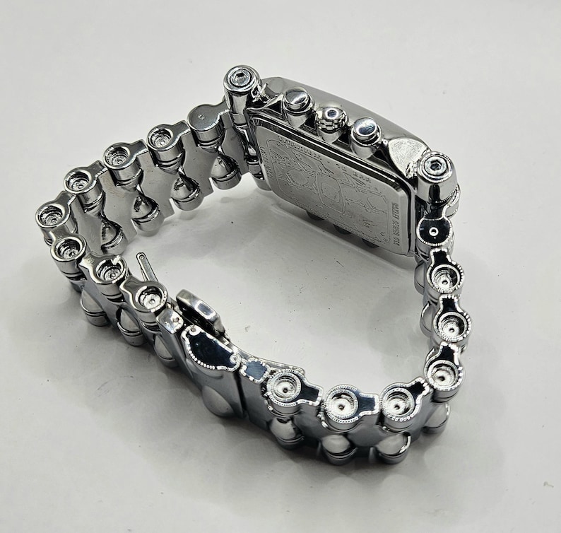 May include: A silver-toned wristwatch with a rectangular face and a chain-link band. The watch face has a detailed design. The band is made of interlocking metal pieces, creating a unique, industrial aesthetic. The watch is shown on a white surface.