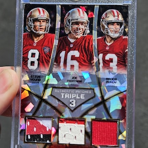 May include: A collectible sports trading card encased in a clear plastic holder featuring three San Francisco 49ers quarterbacks: Steve Young, Joe Montana, and Brock Purdy. The card includes three authentic jersey patches and holographic foil accents.