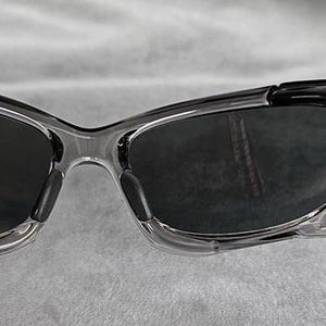 May include: Close-up of a pair of sunglasses with a clear frame and dark lenses. The temples are black and attached to the frame with silver accents. The sunglasses are designed for outdoor wear.