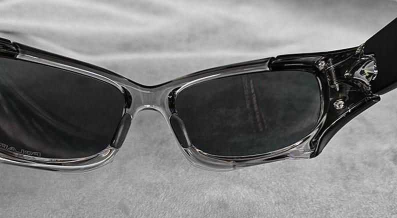 May include: Close-up of a pair of sunglasses with a clear frame and dark lenses. The temples are black and attached to the frame with silver accents. The sunglasses are designed for outdoor wear.