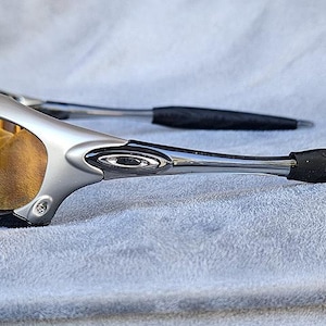 Może przedstawiać: Srebrno-czarne okulary przeciwsłoneczne sportowe ze złotymi soczewkami. Okulary przeciwsłoneczne mają elegancki, nowoczesny design z zakrzywioną oprawką i czarnymi gumowymi końc&oacute;wkami zausznik&oacute;w. Soczewki są w ciepłym, złotym kolorze.